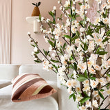 Een gezellige woonkamer met een witte bank, een gestreept roze en bruin kussen en de Kunstplant Kersenbloesem 120 cm crème bloeiende tak op de voorgrond. Een potplant rust op een wandplank tegen een lichtroze muur.