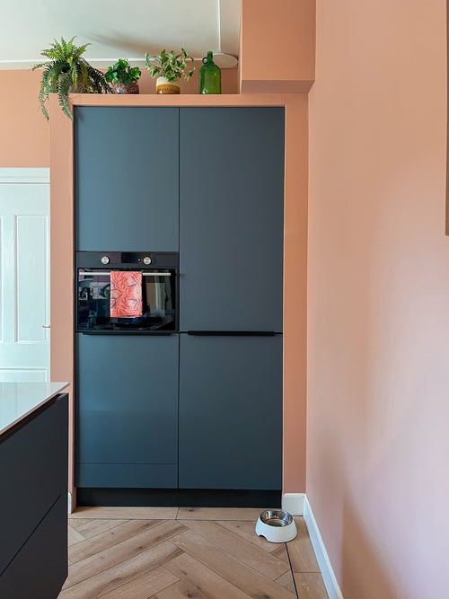 Een moderne keukenhoek met matte donkere kasten, een ingebouwde oven en een lichte houten vloer. De Kunstplant Monstera 18 cm en een groene glazen fles staan bovenop de kastjes; een voerbak voor huisdieren staat op de perzikkleurige muur.