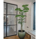 De Kunstplant Monstera 180 cm, een hoge potplant met grote gespleten bladeren, staat in een moderne, lichte kamer naast een glazen deur, houten vloer, bank en blauwe verticale lamellen.