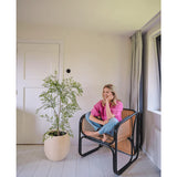 Een vrouw in een roze shirt en spijkerbroek zit blootsvoets op een geweven stoel, glimlachend en ontspannend in een lichte, minimalistische kamer met witte muren en vloeren. Naast haar staat de Kunstplant Ruscus 120 cm.