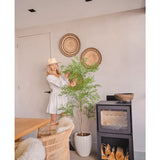 Een vrouw in een witte jurk en hoed staat naast de Kunstplant Ruscus 150 cm, die een natuurlijke flair toevoegt aan een moderne kamer met rieten meubels, muurmanden en een zwarte houtkachel - perfect voor decoratieve interieurs.