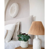 Een moderne slaapkamer met een neutraal kleurenpalet heeft een bed met wit beddengoed, een houten nachtkastje, een lamp van gestructureerd keramiek met geweven kap, de Kunstplant Saxifraga 27 cm, en een ronde witte wandsculptuur boven het hoofdeinde.