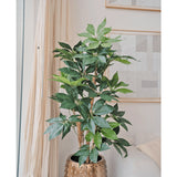De Kunstplant Schefflera 150 cm met brede groene bladeren staat in een goudkleurige pot naast een crèmekleurige bank en lichte gordijnen, terwijl aan de muur abstracte kunst hangt.