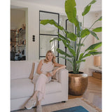 Een vrouw in lichte kleding zit met een mok op een beige bank. Naast haar brengt de Kunstplant Strelitzia Nicolai Deluxe 235 cm weelderig groen in de stijlvolle, moderne woonkamer met neutrale inrichting.