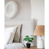 Een gezellige slaapkamer heeft een bed met neutraal beddengoed, een licht houten nachtkastje met de Kunstplant Watermeloen 29 cm en een witte lamp met een rieten kap. Boven het bed hangt een witte wandsculptuur met textuur.