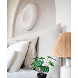 Een keurig opgemaakt bed met witte kussens en beddengoed staat naast een houten nachtkastje met de Kunstplant Watermeloen 29 cm en een lamp met een geweven kap; boven het hoofdeinde hangt een wandsculptuur met witte textuur.
