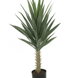 De Kunstplant Yucca 100 cm UV heeft hoge, stekelige groene bladeren op een dikke stengel, is opgepot in een kleine zwarte pot met aarde en is gefotografeerd tegen een witte achtergrond.