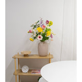 Een beige vaas met Kunstranonkel Real Touch Geel 60cm bloemen staat op een lichte houten plank naast een ronde witte tafel. Op de plank staan ook een kleine witte klok, een houten dienblad en een boek tegen een effen witte muur.