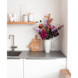 Een moderne witte keuken met een grijs aanrechtblad, een gootsteen, een houten snijplank en een witte vaas gevuld met Kunstbloem Hortensia 55 cm donkerroze. Witte kopjes en potten staan op een houten plank boven het aanrecht.