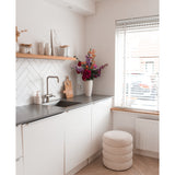 Een moderne, minimale keuken met witte kastjes, een zwart aanrechtblad en een Kunstbloem Hortensia 55 cm donkerroze in een vaas bij het aanrecht, plus houten planken met decor en een ronde kruk met kussens bij een raam met jaloezieën.