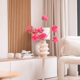 Een moderne woonkamer is voorzien van een witte bijzettafel met de Kunsttak Kersenbloesem 85 cm roze, waaraan felroze bloemen zijn toegevoegd. Een neutrale inrichting, lichte houtaccenten, beige gordijnen en een fauteuil met structuur maken de gezellige, minimalistische ruimte compleet.