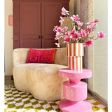 Een gezellige woonkamer met een beige ronde bank, een roze en oranje geometrisch kussen, een roze bijzettafel, Kunsttak Magnolia kleinbloemig 89 cm roze in een vaas, een geel-wit gedessineerd vloerkleed en een kast in kastanjebruin op de achtergrond.
