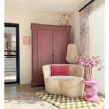 Een gezellige leeshoek heeft een crèmekleurige stoel, roze kussens, een geel gedessineerd vloerkleed en een mauve kast. Op een roze bijzettafel staat de Kunsttak Magnolia kleinbloemig 89 cm roze, terwijl grote ramen deze uitnodigende ruimte vullen met zacht licht.