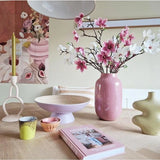Een moderne kamer heeft een gedekte tafel met Kunsttak Magnolia kleinbloemig 89 cm roze in een roze vaas, pastelkleurige kandelaars, schalen, kopjes, een groene sculpturale vaas, een roze boek en bloemenkunstwerk aan de muur.