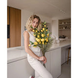 Een vrouw met krullend blond haar, gekleed in een crèmekleurige outfit, leunt tegen een aanrecht en houdt het boeket ‘Kunsttak Lemon 104 cm geel’, versierd met weelderig groen, vast. De moderne keuken is uitgevoerd in neutrale kleuren en heeft houten accenten.