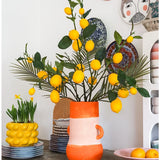 Een lichte kamer met een hoge vaas vol citroenen en groene bladeren, waarin de opvallende Kunsttak Lemon 104 cm geel een prominente plaats inneemt in het arrangement, omringd door kleurrijke keramische schalen, een schaal met citroenen en decoratieve borden aan de muur – wat een vrolijke, eclectische uitstraling creëert.