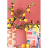 Tegen een koraalroze muur staan een gestreepte vaas gevuld met Kunsttak Lemon-takken van 104 cm in geel, een kleurrijk schilderij, een stapel boeken, een blauwe kandelaar met een kaars en een dienblad met citroenen.