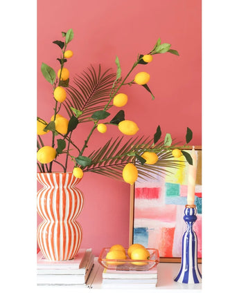 Tegen een koraalroze muur staan een gestreepte vaas gevuld met Kunsttak Lemon-takken van 104 cm in geel, een kleurrijk schilderij, een stapel boeken, een blauwe kandelaar met een kaars en een dienblad met citroenen.