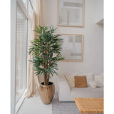 Een Lady Kunstpalm 180 cm is geplaatst bij een groot raam in een lichte, moderne woonkamer met een witte bank, beige kussens, geweven tafel en abstracte kunst. Het voegt een gezellige minimalistische stijl toe met zijn neutrale tinten en groene kunstplant charme.