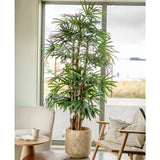 Een 210 cm hoge Lady Kunstpalm staat naast een raam in een lichte, moderne woonkamer met een stoel met kussens, een kleine ronde tafel en een neutrale inrichting.
