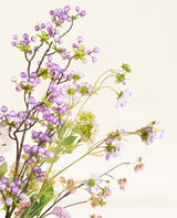 Het Zijden kunstbloemen boeket Lilac heeft delicate kunstmatige paarse bessen, kleine lila bloesems, witte bloemen en groene bladeren tegen een lichtbeige achtergrond.