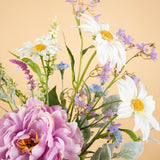 Het Zijden kunstbloemen boeket Misty Minthe heeft witte margrieten, paarse en blauwe bloesems en een grote roze pioenroos tegen een zacht perzikkleurige achtergrond.