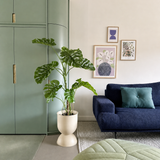 Een moderne woonkamer met een groene inbouwkast, Kunstplant Monstera 120 cm, een blauwe bank met een groenblauw kussen, abstracte kunst aan de muur en een groene poef op een licht vloerkleed.