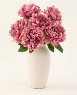 De Zijden kunstbloemen boeket Peony Dusty Pink is gevat in een witte vaas van keramiek met textuur. De vaas heeft grote roze pioenrozen met weelderige groene bladeren tegen een lichte, effen achtergrond.