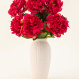 Een vaas van wit keramiek met textuur bevat zes grote, levendige zijden bloemen uit het Zijden kunstbloemen boeket Peony Raspberry Red, met weelderige groene bladeren, tegen een effen lichte achtergrond.