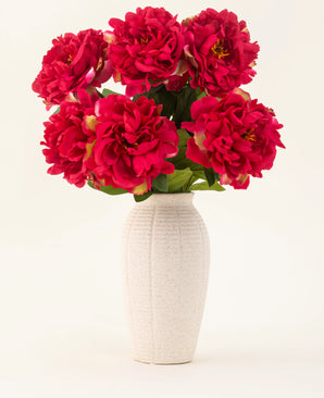 Een vaas van wit keramiek met textuur bevat zes grote, levendige zijden bloemen uit het Zijden kunstbloemen boeket Peony Raspberry Red, met weelderige groene bladeren, tegen een effen lichte achtergrond.