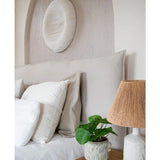 Een knusse slaaphoek met een beige hoofdeinde, witte en beige kussens, een houten nachtkastje, een keramische lamp met een geweven kap, de Kunst Pannenkoekplant 27 cm en witte muurkunst boven het bed.