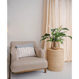 Een beige fauteuil met een decoratief kussen staat naast een ronde rieten bijzettafel met daarop de Kunstplant Spathiphyllum wit 85 cm. Lichte gordijnen op de achtergrond maken dit rustige, moderne interieur compleet.