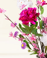 Het Zijden kunstbloemen boeket Pink Reverie heeft een grote magenta pioenroos, kleine roze bloemen, paarse madeliefachtige bloemen en groene bladeren in een vaas-een romantisch boeket tegen een lichte achtergrond.