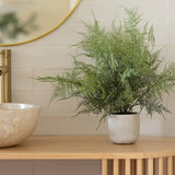 De Kunstplant Asparagus 43 cm in sierpot staat op een houten aanrecht naast een ronde stenen wastafel en gouden kraan, met een ronde spiegel die het tafereel weerspiegelt tegen een wit betegelde muur.
