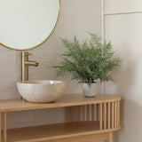 Een minimalistisch badkamermeubel heeft een ronde spiegel, een gouden kraan, een stenen wasbak en de Kunstplant Asparagus 43 cm in sierpot op een licht houten aanrechtblad tegen wit betegelde muren.