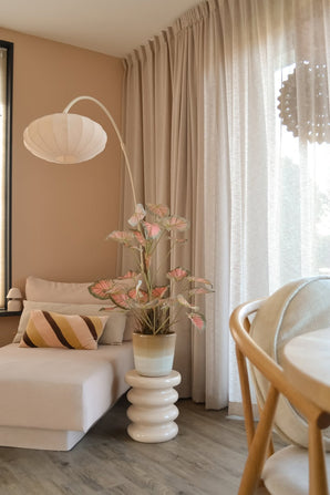 Een gezellige zithoek met een beige chaise longue, een gestreept kussen, de Kunstplant Caladium 90 cm roze die voor een vleugje groen zorgt, een moderne lamp, lichte gordijnen en een houten vloer. Zacht natuurlijk licht valt door de transparante gordijnen naar binnen.