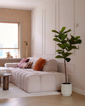Een moderne woonkamer met een pluche lichtbeige bank, kleurrijke kussens, de Kunstplant Ficus Tabaksplant Deluxe 155 cm in de hoek, een kleine ronde tafel en jaloezieën. Warme neutrale tinten en structuurbehang creëren een gezellige sfeer.
