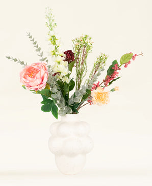 Een witte, gestructureerde vaas met een ronde, sprankelende vorm toont het Zijden kunstbloemen boeket Pretty Petite-roze, gele en witte bloemen met groen en rood gebladerte-ideaal voor een duurzaam interieur tegen een effen lichte achtergrond.