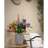 Een witte vaas met vijf levendige oranje Ranonkel kunsttak x5 Real Touch 62cm bloemen staat op een houten tafel naast een houten stoel met een wit kussen en een deken; gevlochten manden hangen aan een beige muur en groene takken gluren dichtbij.