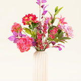 Een crèmekleurige, geribbelde vaas van keramiek bevat het Zijden kunstbloemen boeket Rosy Saga Mist, met roze, rode en paarse zijden bloemen met groene bladeren tegen een lichte achtergrond.