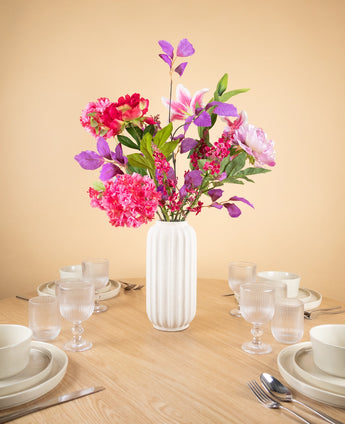 Een witte vaas van keramiek met het Zijden kunstbloemen boeket Rosy Saga Mist fleurt een houten eettafel op voor vier personen met witte borden, kommen, kopjes en glazen, tegen een effen beige muur.