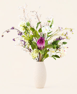Een hoge, witte vaas met textuur bevat het Zijden kunstbloemen boeket Serene Bloomfield, met een grote paarse protea, witte en paarse bloemen, groene bladeren en sierlijke takken - perfect voor een elegante touch in huis.