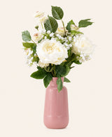 Het Zijden kunstbloemen boeket White Love heeft witte pioenrozen, kleine witte vulbloemen en groene bladeren in een roze geribbelde vaas van keramiek, tegen een effen gebroken witte achtergrond.