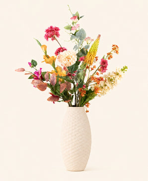 Een crèmekleurige vaas met structuur bevat het Zijden kunstbloemen boeket Wild Peach, met roze, gele, oranje en witte bloemen met groen gebladerte, tegen een effen lichte achtergrond.