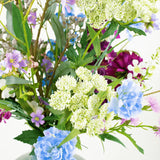 Het zijden kunstbloemen boeket Purple Rain met lichtgroene Queen Anne's lace, blauwe anjers, paarse en roze bloemen in een groene vaas toont Nederlands vakmanschap tegen een witte achtergrond.