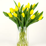 Het Zijden kunstboeket Tulpen in vaas Geel bestaat uit een boeket gele tulpen met groene bladeren in een heldere glazen vaas, tegen een effen witte achtergrond.