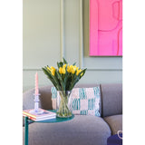 Het Zijden kunstboeket Tulpen in vaas Geel staat op een groen bijzettafeltje naast een roze gedraaide kaars, twee boeken en een grijze gestructureerde bank met een kussen met patroon. Aan de lichtgroene muur hangt een felroze abstract kunstwerk.