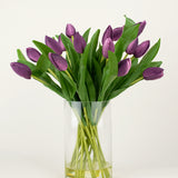 Het Zijden kunstboeket Tulpen in vaas Paars heeft langstelige paarse kunsttulpen met weelderige groene bladeren, prachtig gerangschikt in een heldere glazen vaas tegen een effen witte achtergrond.