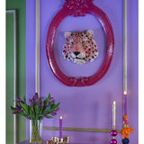 Een levendige paarse muur toont een decoratieve luipaardkop in een roze sierlijke lijst. Hieronder zie je het Zijden kunstboeket Tulpen in vaas Paars, naast een paarse kaars, een gouden kandelaar en kleurrijke kaarsen op een plank.