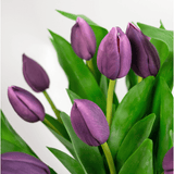 Een close-up van het Zijden kunstboeket Tulpen in vaas Paars toont een aantal paarse tulpenbloemen met groene bladeren in een vaas tegen een lichte, neutrale achtergrond.